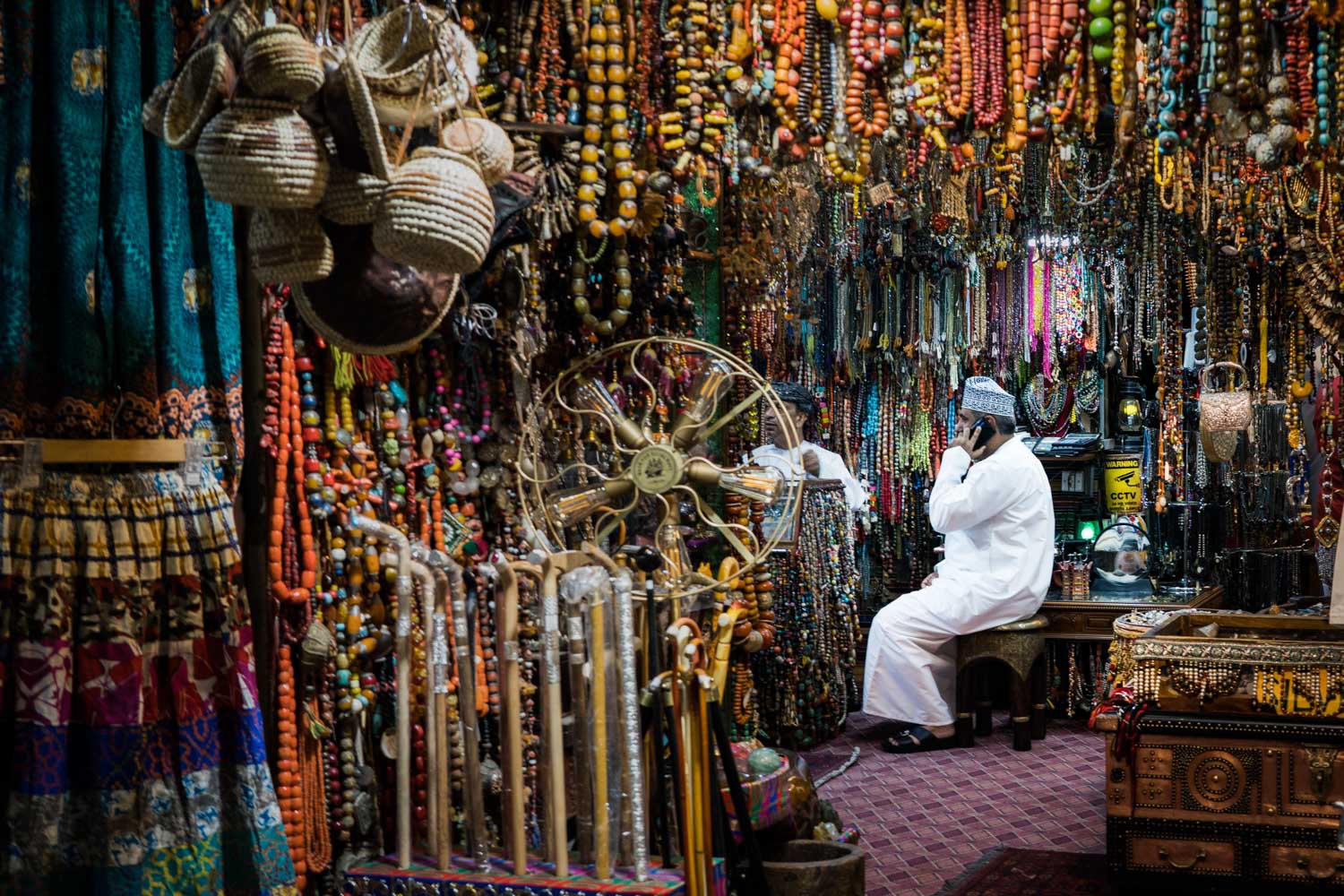 Man in traditional attire on the phone in a vibrant market stall filled with beads, textiles, and baskets.