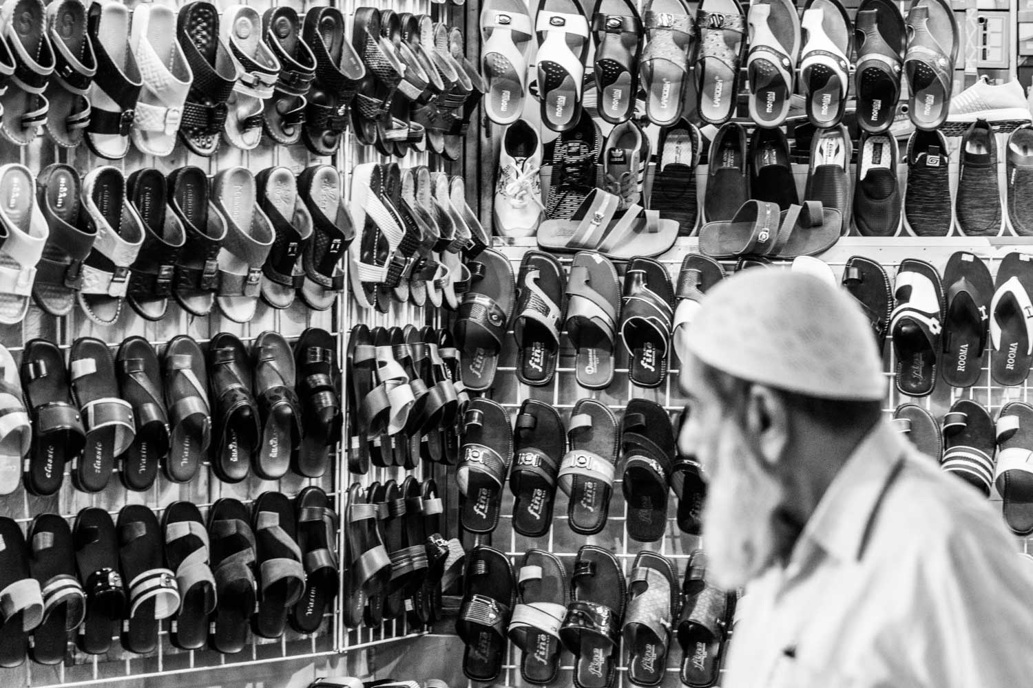 Black and white image of numerous sandals displayed on a wall, with a man in profile wearing a cap in the foreground.