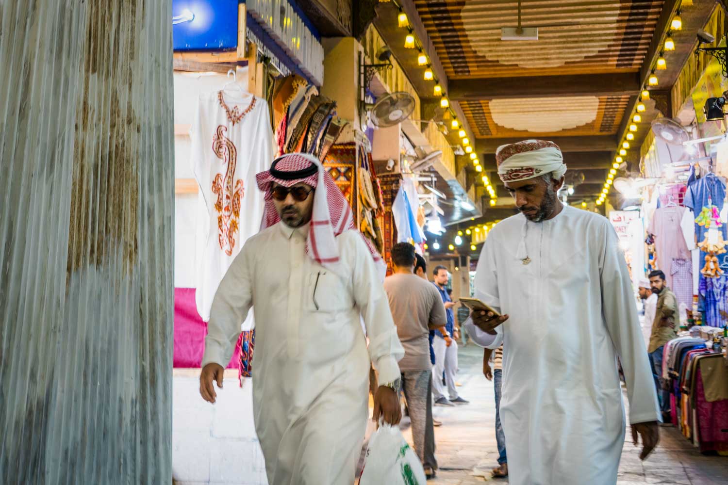 Men in traditional clothing walk through a vibrant Middle Eastern market, lined with colorful textiles and decorations.