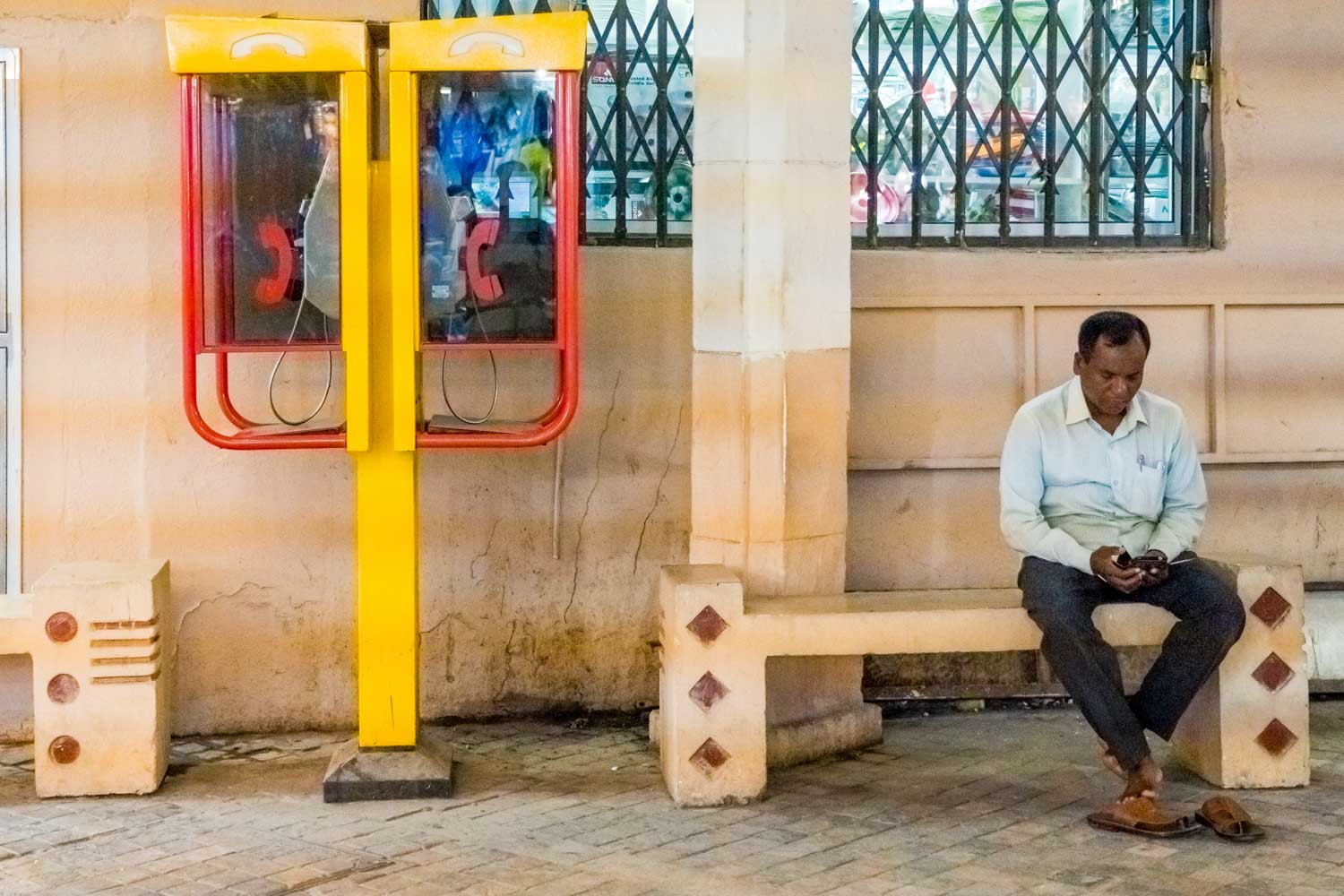 Man sitting on a bench, using a smartphone next to two yellow and red payphones on a wall with barred window.