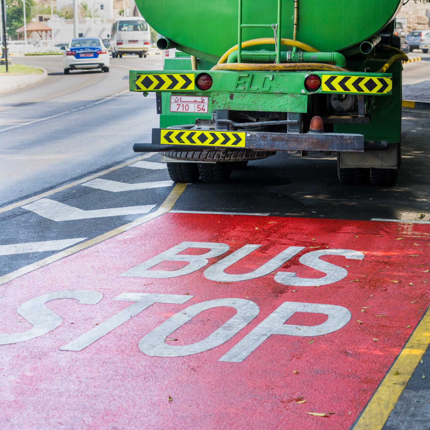 Green truck parked in bus stop zone on street with red markings and traffic in the background.