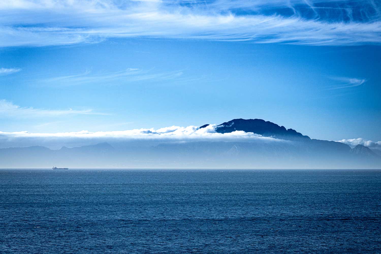 Vast ocean view with a distant ship and a mountain range under a clear blue sky and scattered clouds.