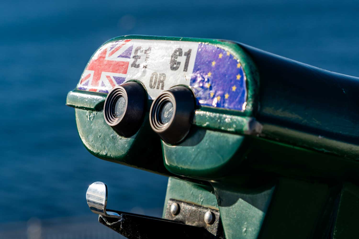 Coin-operated binoculars with UK and EU flags, overlooking the sea on a sunny day.