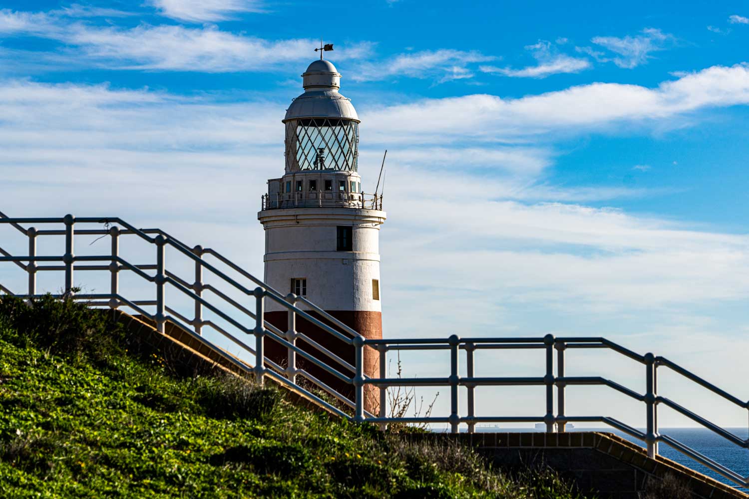 Lighthouse with a metal railing in foreground, blue sky, and ocean horizon in the background.