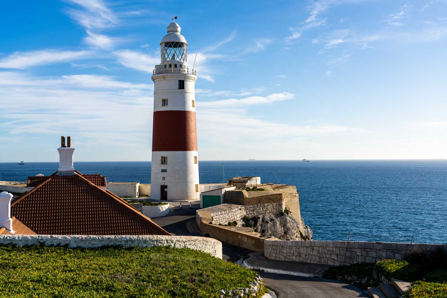 Lighthouse by the sea with a clear blue sky, near a building with a red-tiled roof, surrounded by ocean views.