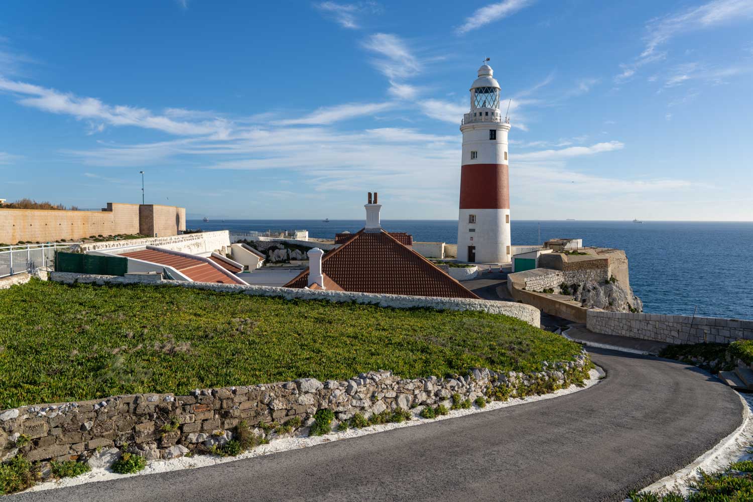 Lighthouse near the sea on a sunny day, with green grass and a winding road in the foreground.