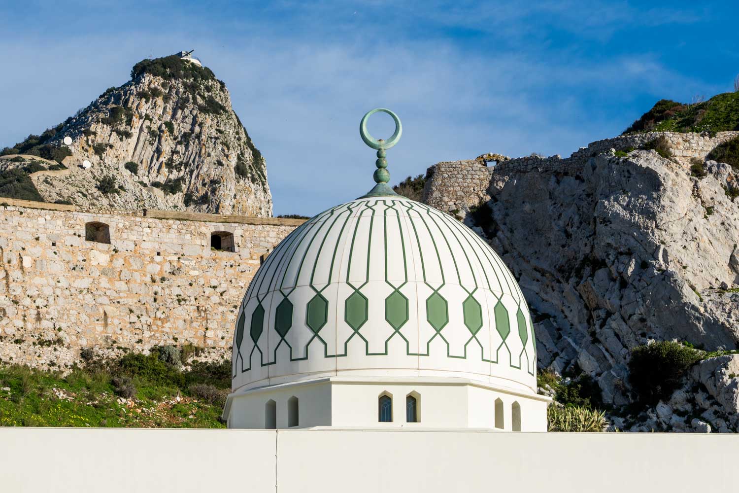 White mosque dome with green details against rocky hillside in Gibraltar, under a clear blue sky.