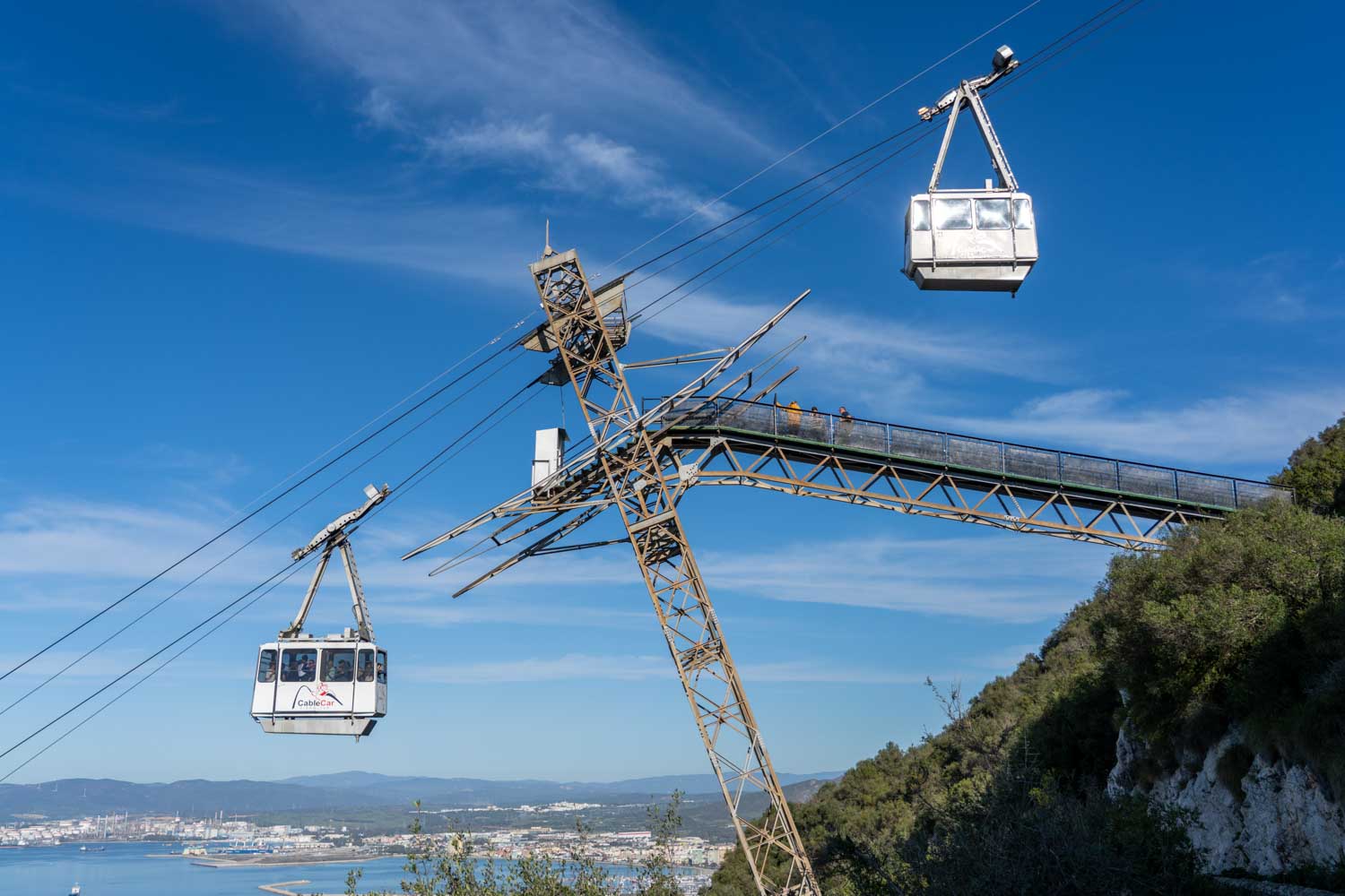 Cable cars ascending over hillside with platform, blue sky backdrop, and coastal city view below.