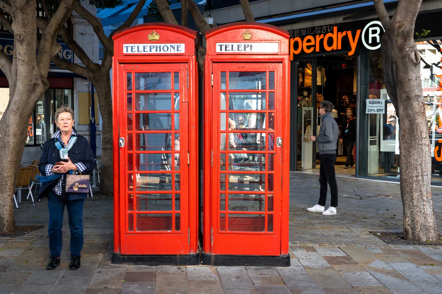 Red telephone booths beside a Superdry store, with people walking on a city street.