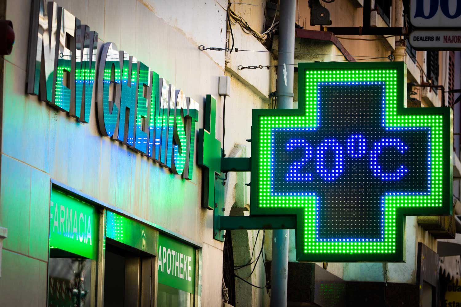 Pharmacy sign with a green cross and LED display showing 20°C, next to NEW CHEMIST in various languages.