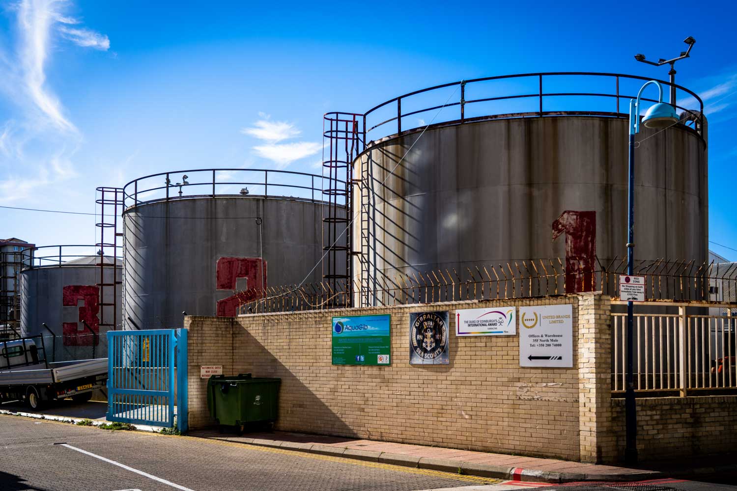 Large industrial tanks numbered 1, 2, and 3 behind a brick wall and blue gate under a clear sky.