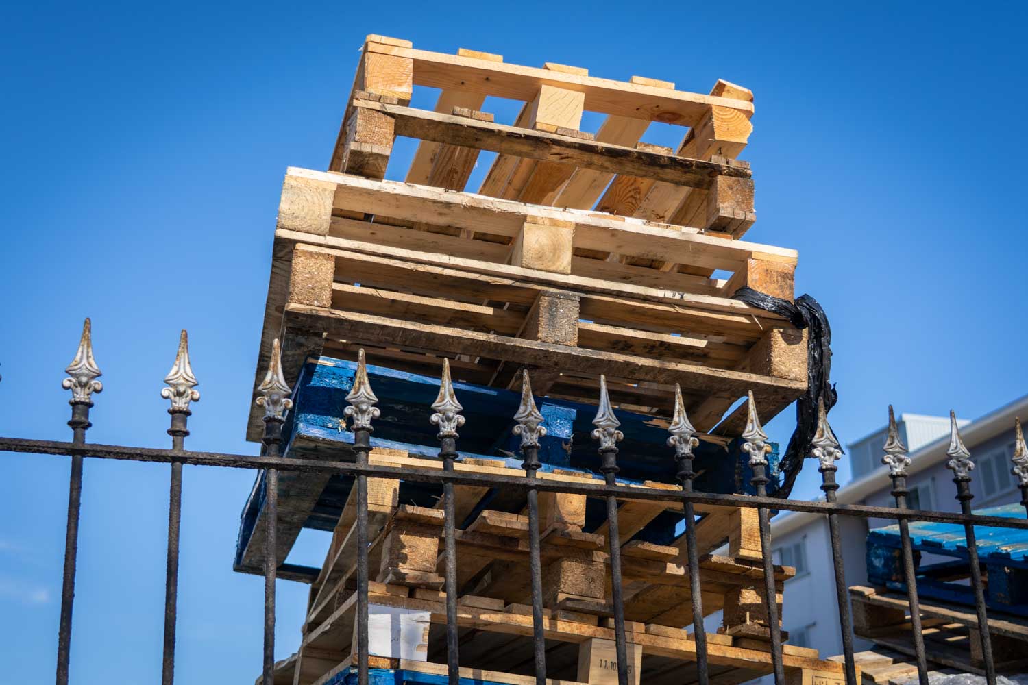 Stacked wooden pallets with blue accents behind a decorative iron fence against a clear blue sky.