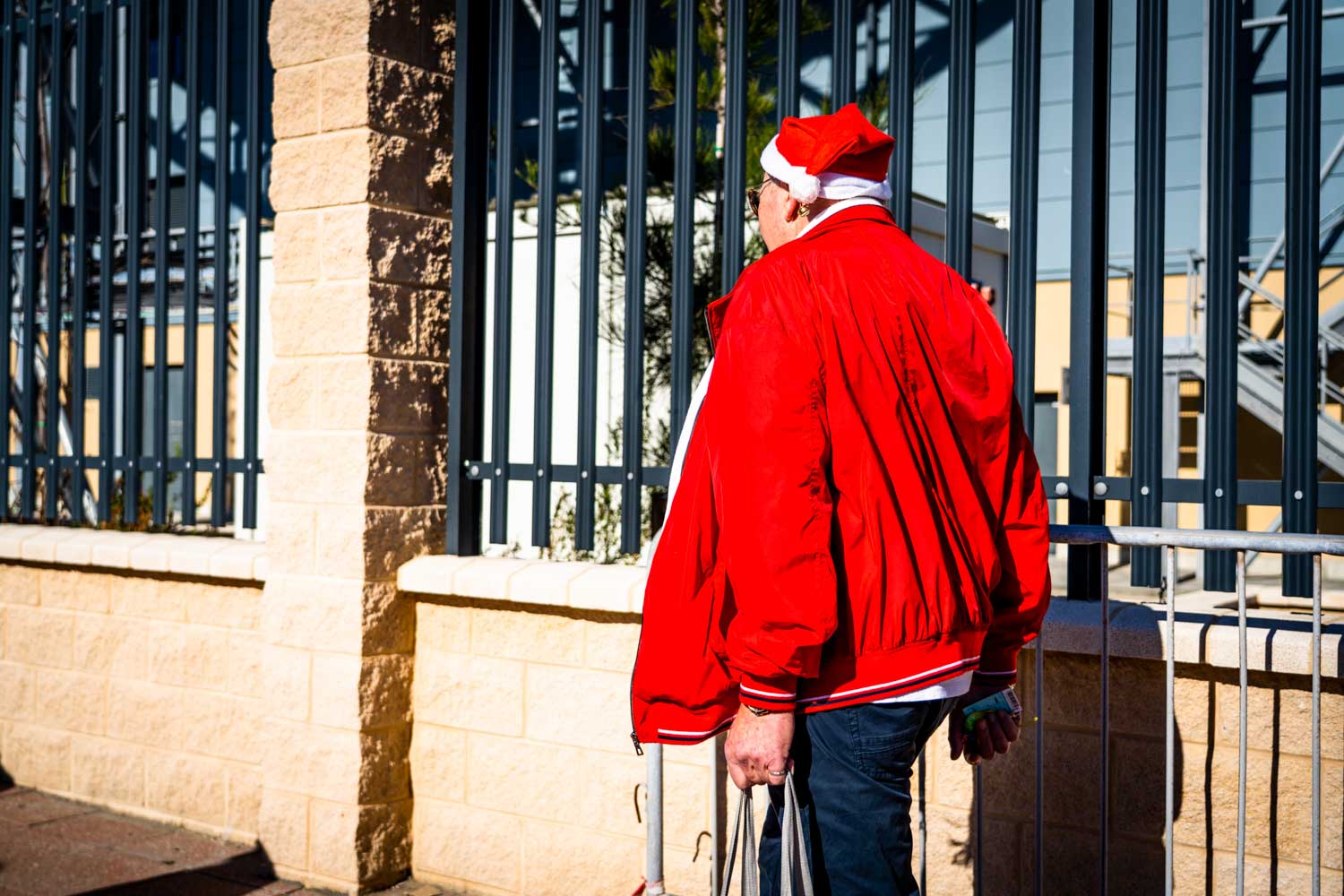 Man in red jacket and Santa hat walking by a metal fence on a sunny day.
