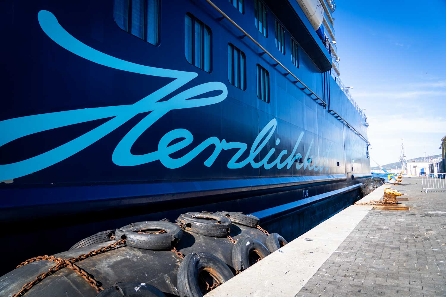Blue cruise ship docked at a port, displaying elegant script lettering, under a clear blue sky.