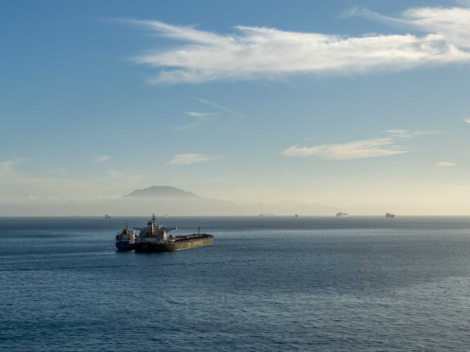 Cargo ship sailing on calm ocean with distant ships and mountain under clear blue sky.