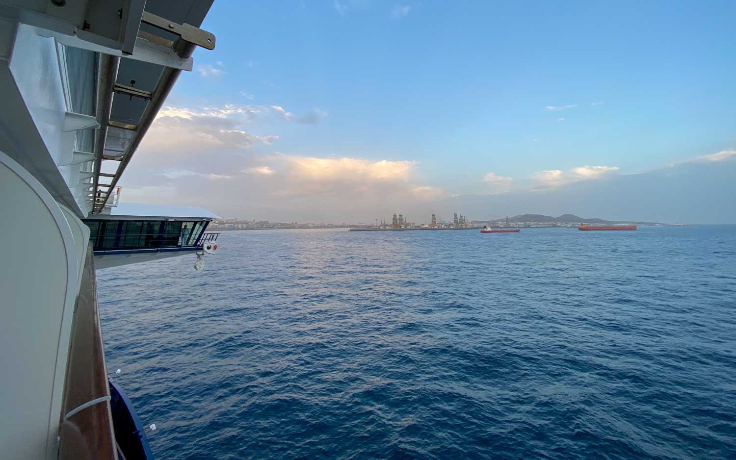View from a ship looking out to sea, with distant cargo ships and an industrial port under a partly cloudy blue sky.