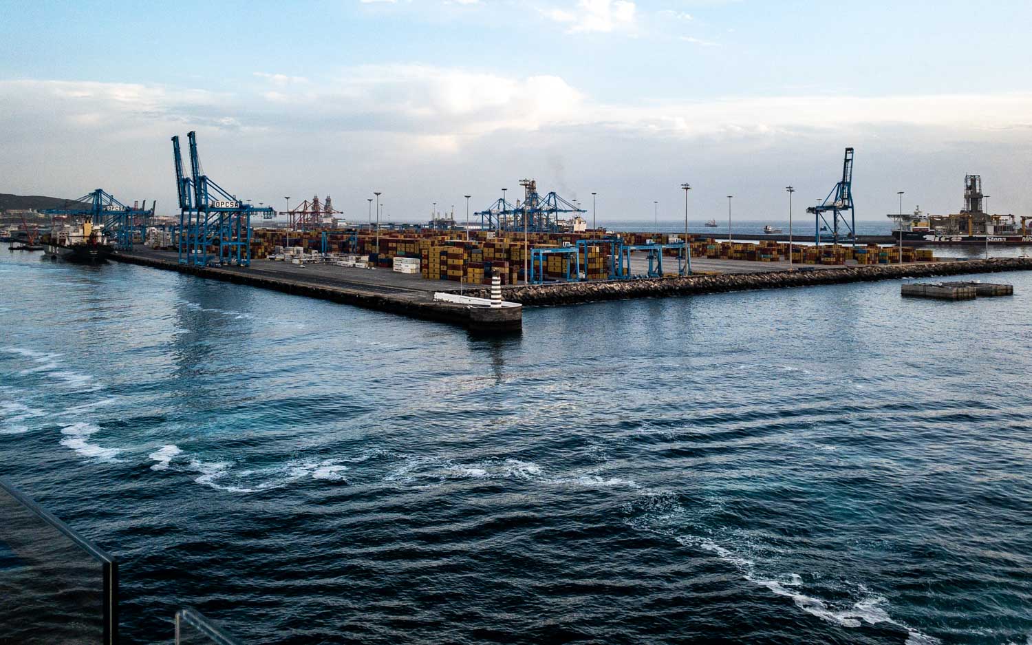 Container port with cranes and stacked cargo near the ocean under a cloudy sky.