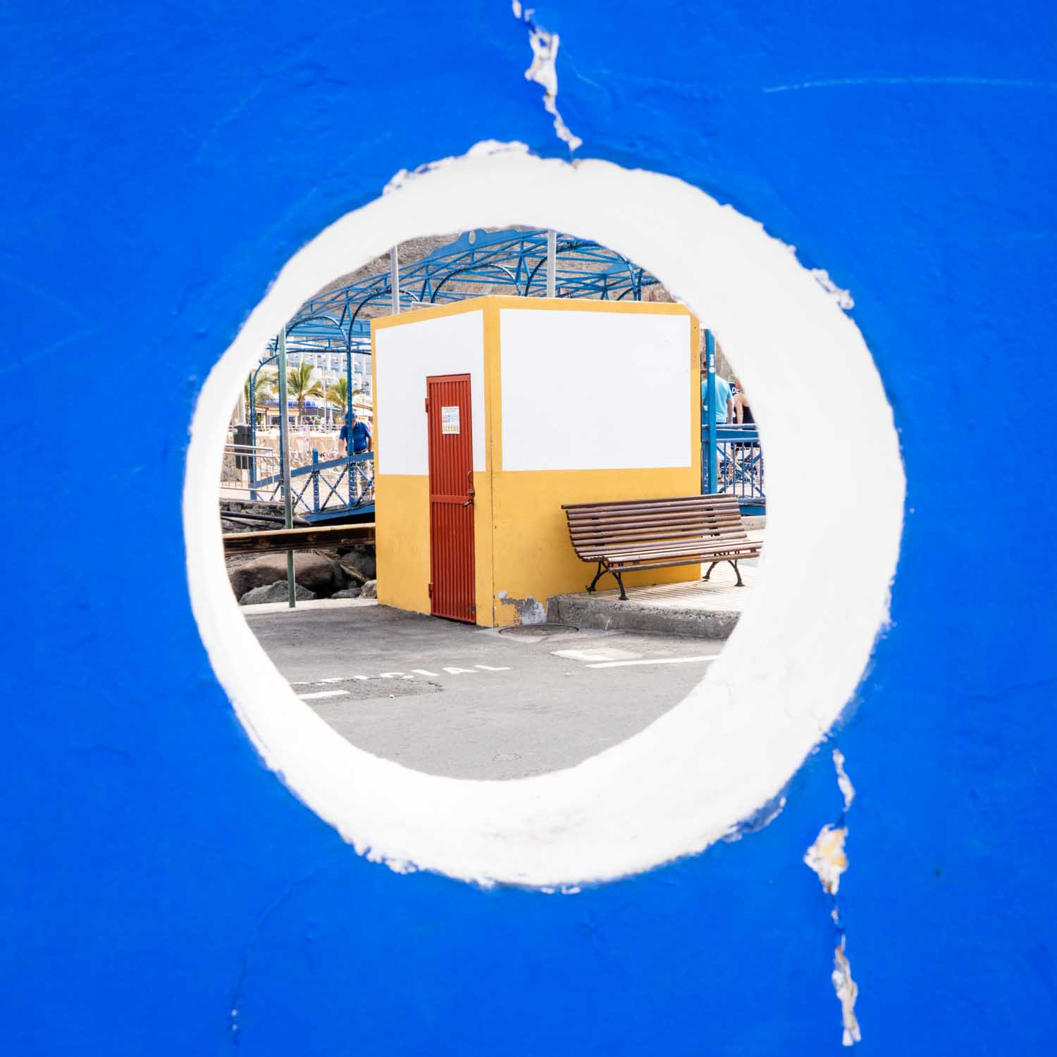 Circular view of a blue wall framing a bench, colorful building, and outdoor area.
