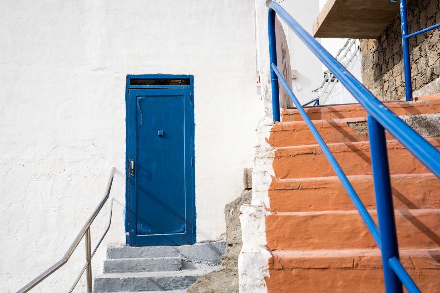 Blue door with white wall, orange stairs, and blue railing in a sunlit coastal setting.