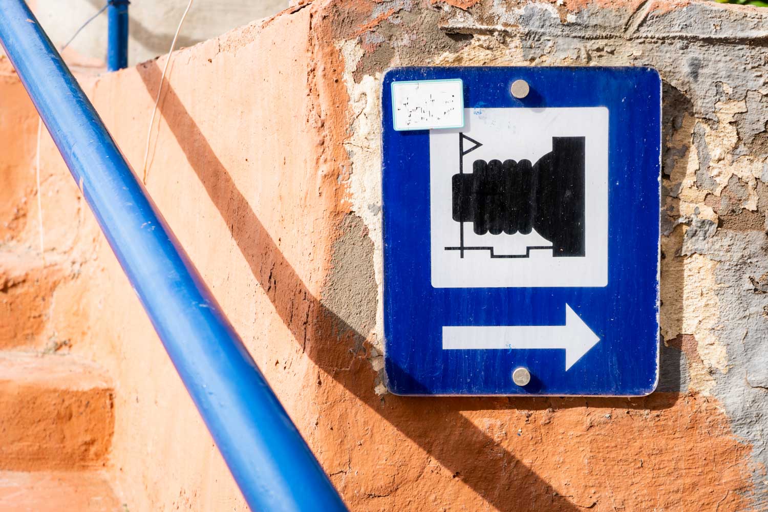 Blue sign on a weathered wall with a camera icon and arrow pointing right, beside blue railing on sandy-colored steps.
