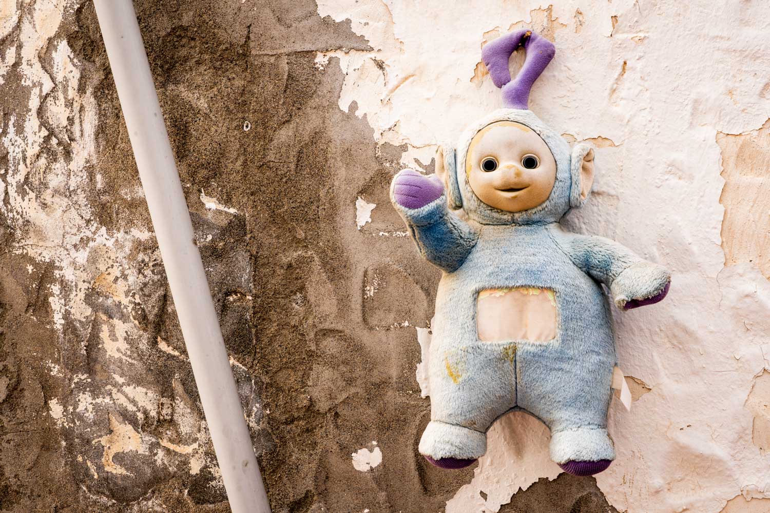 Worn plush toy hanging on a weathered wall with peeling paint, featuring a pipe running alongside.