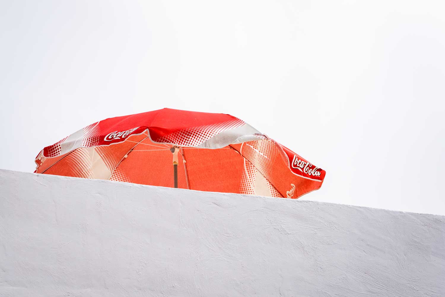 Red Coca-Cola umbrella peeks over white wall against clear sky.