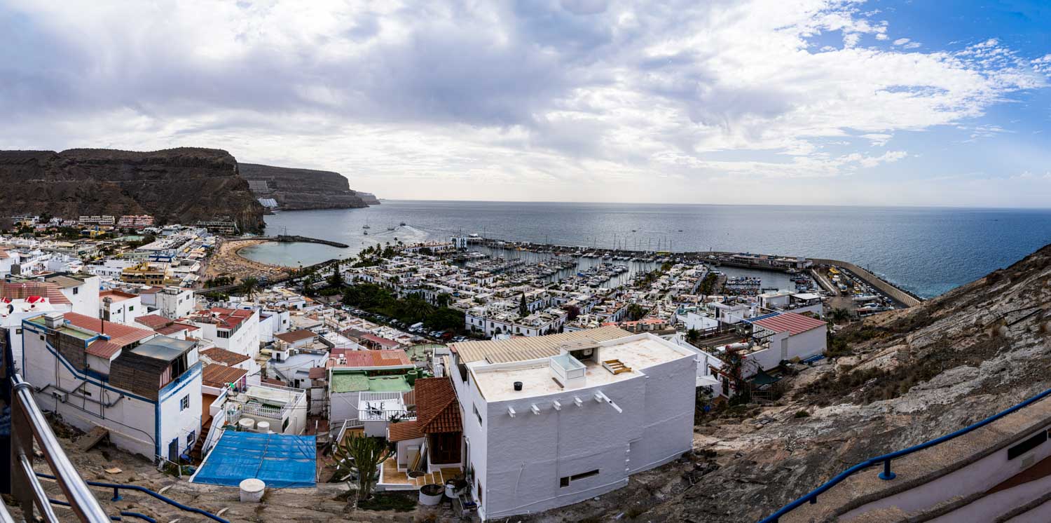Scenic view of coastal town with white buildings, marina, and beach by rocky cliffs under a partly cloudy sky.