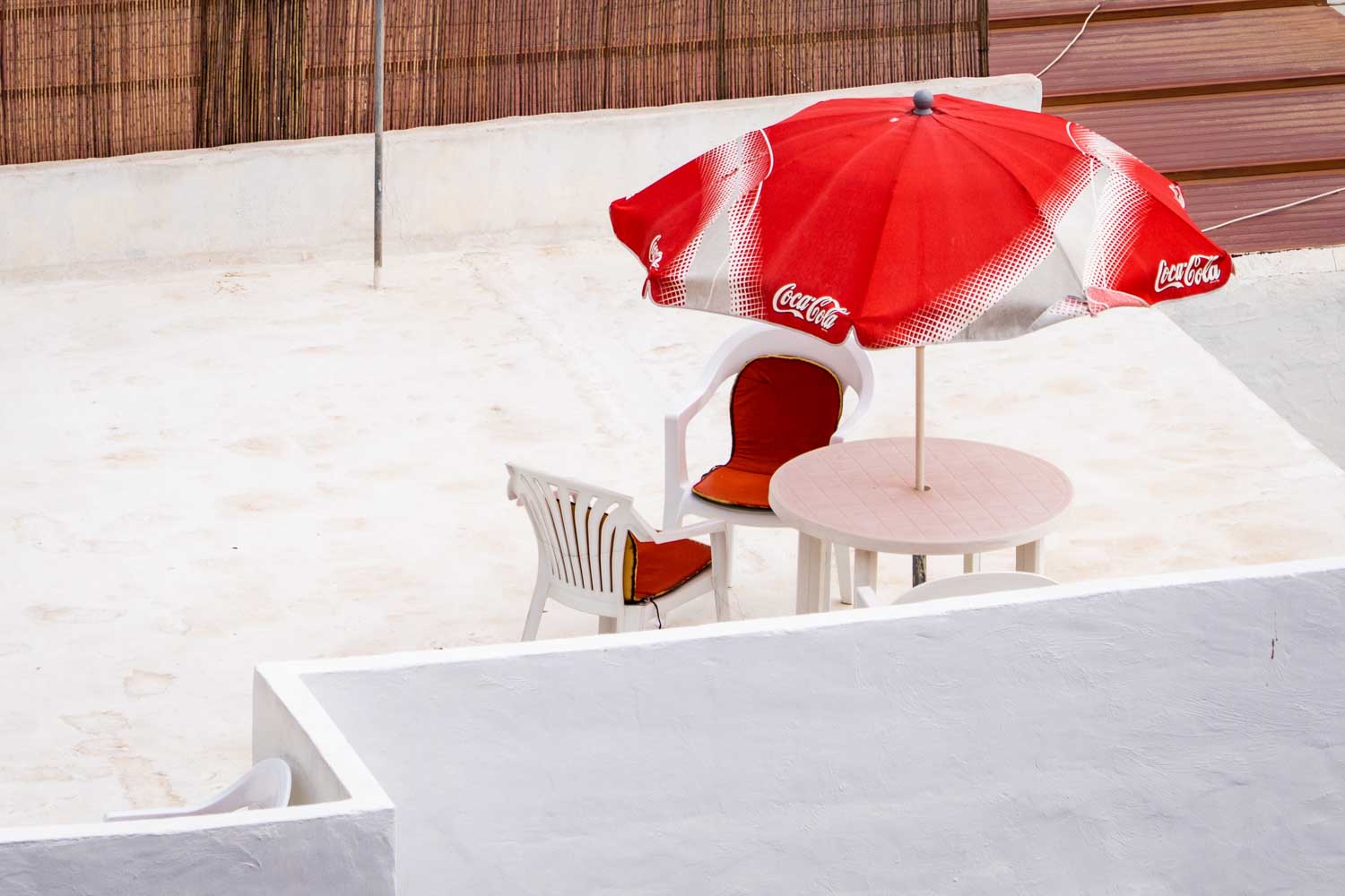 Red Coca-Cola umbrella over white patio chairs and table on a sunny rooftop terrace.