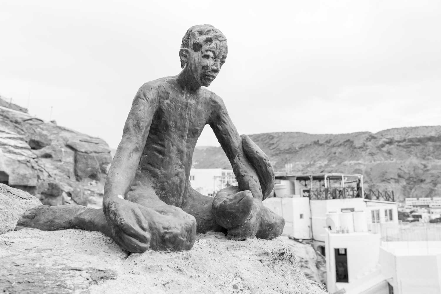Bronze statue of a seated figure on a rocky hillside, with buildings and mountains in the background, in black and white.