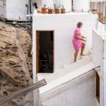 Woman in pink dress climbing white steps beside rocky wall, holding items under clear sky.
