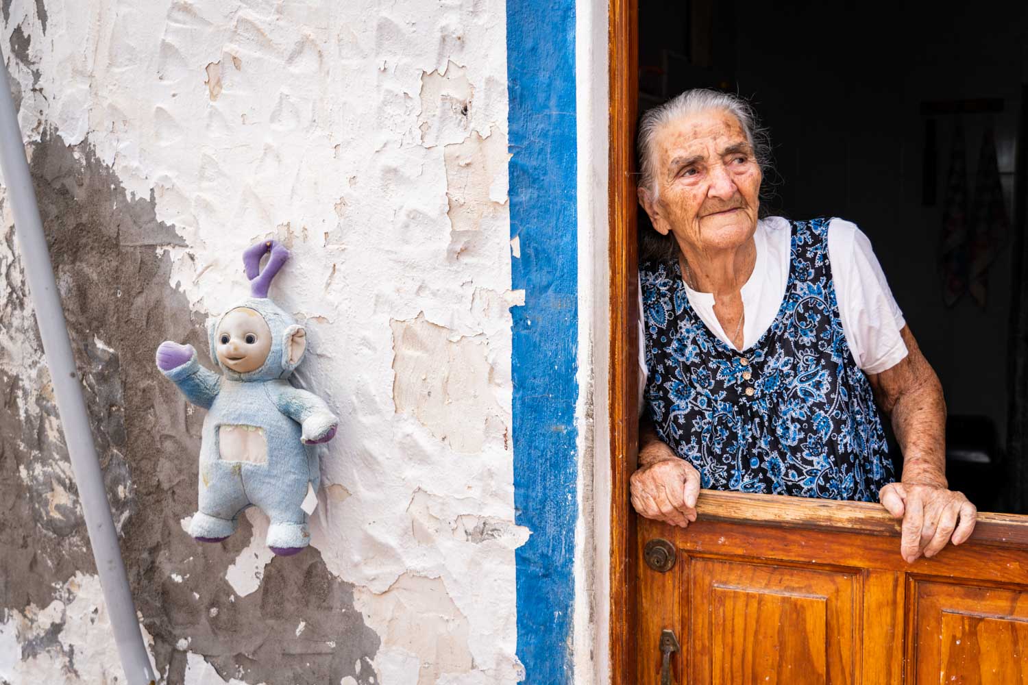 Elderly woman in floral vest leans on wooden door, with a worn plush toy on textured wall beside her.