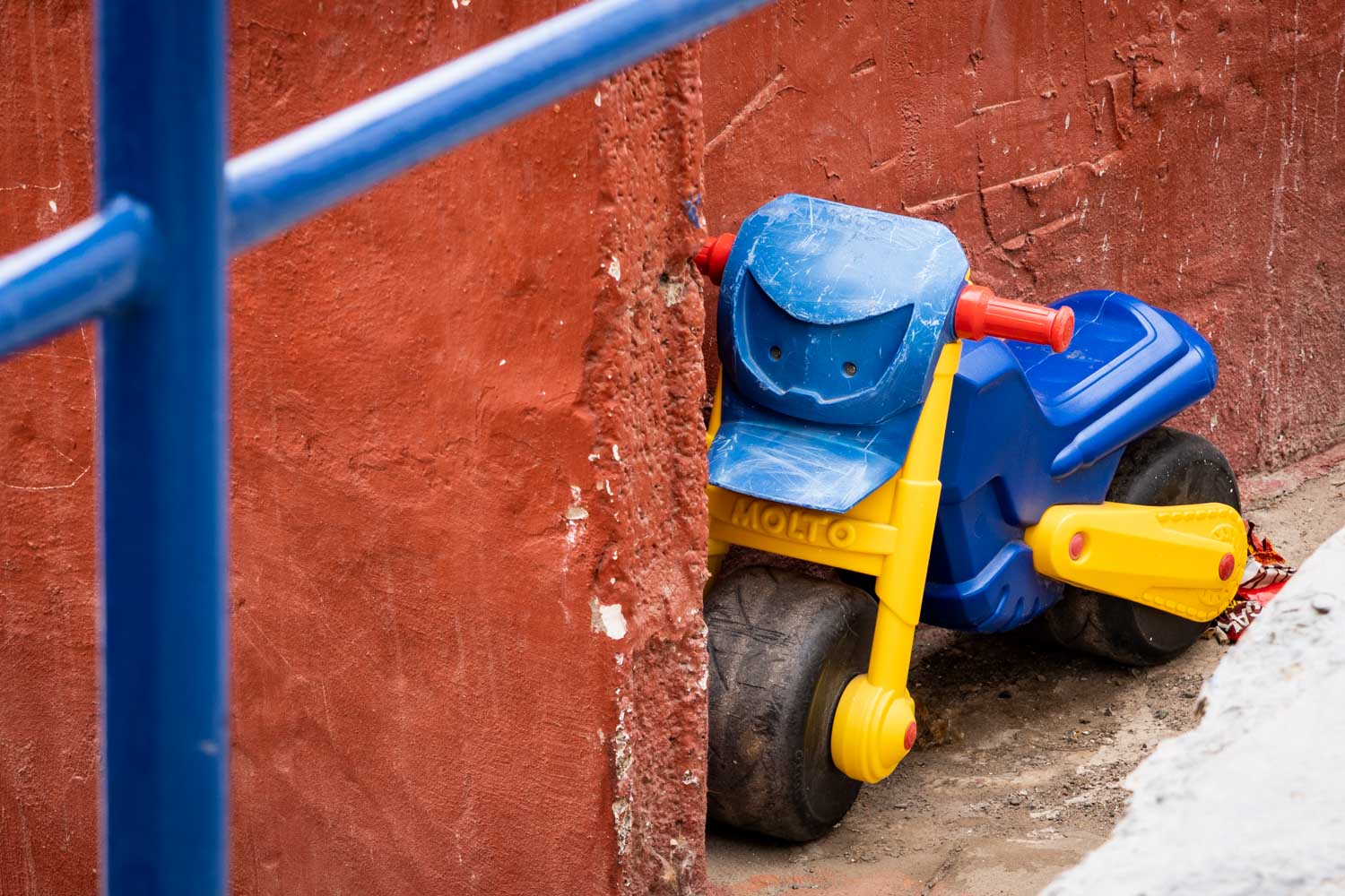 Toy tricycle in blue, yellow, and red abandoned against a red wall with blue railings in the foreground.