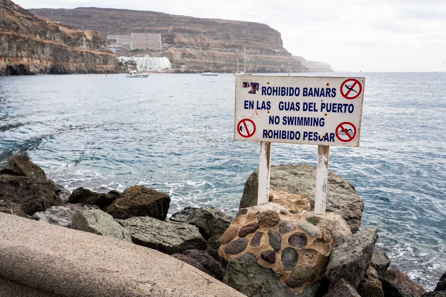 Sign on rocky coast warns against swimming and fishing; ocean and cliffs visible in background.