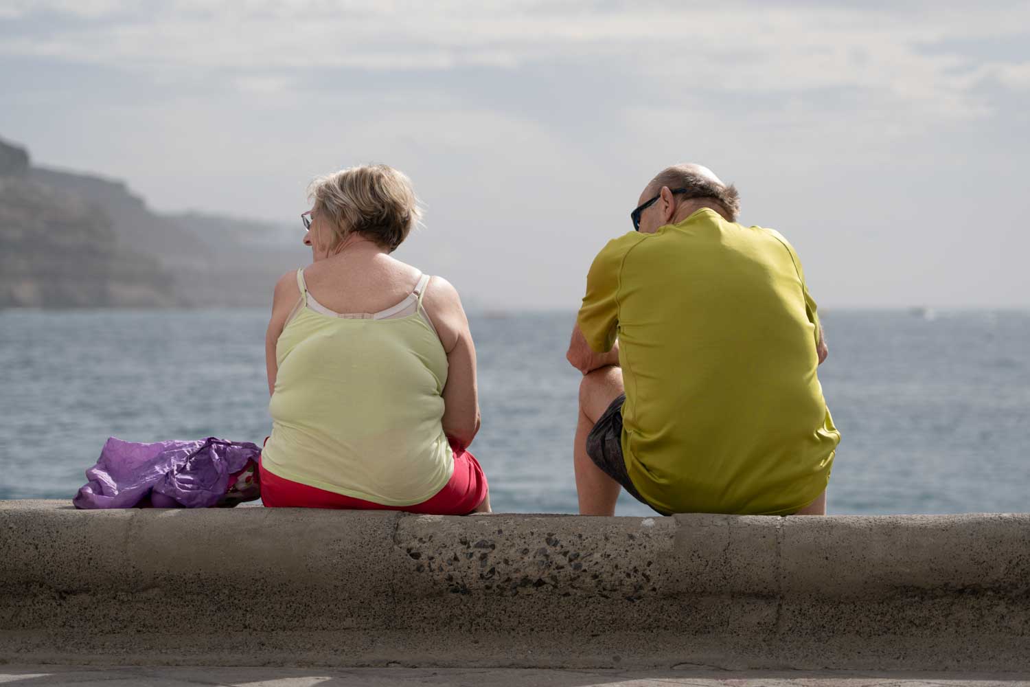 Elderly couple sitting on a concrete wall, overlooking the ocean on a cloudy day.