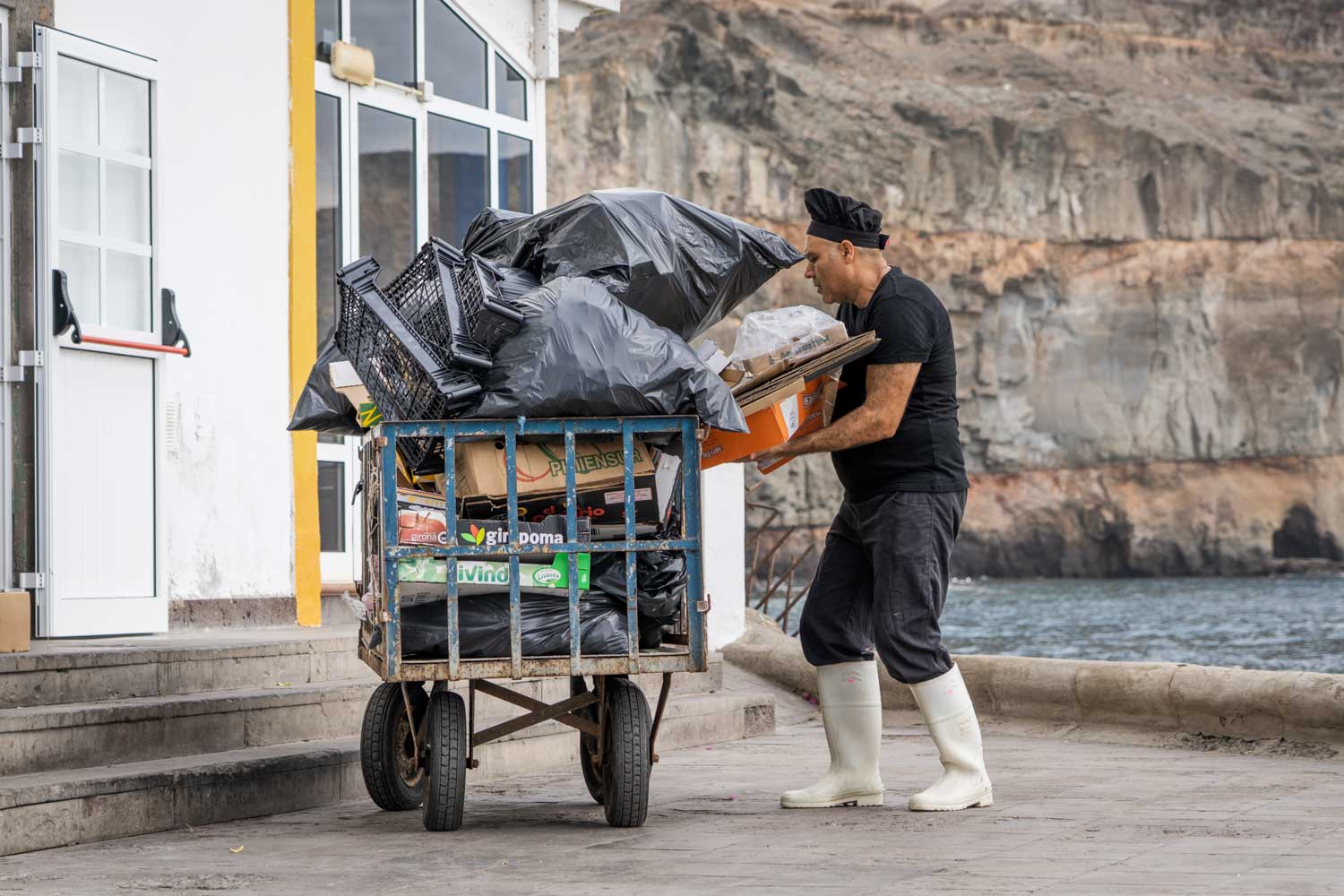 Man in white boots unloading garbage from a cart by the sea near a building.