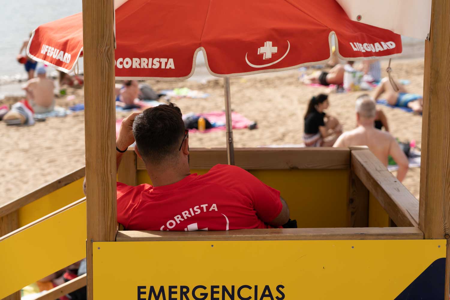 Lifeguard in red shirt monitors beachgoers from wooden lifeguard stand with red umbrella.