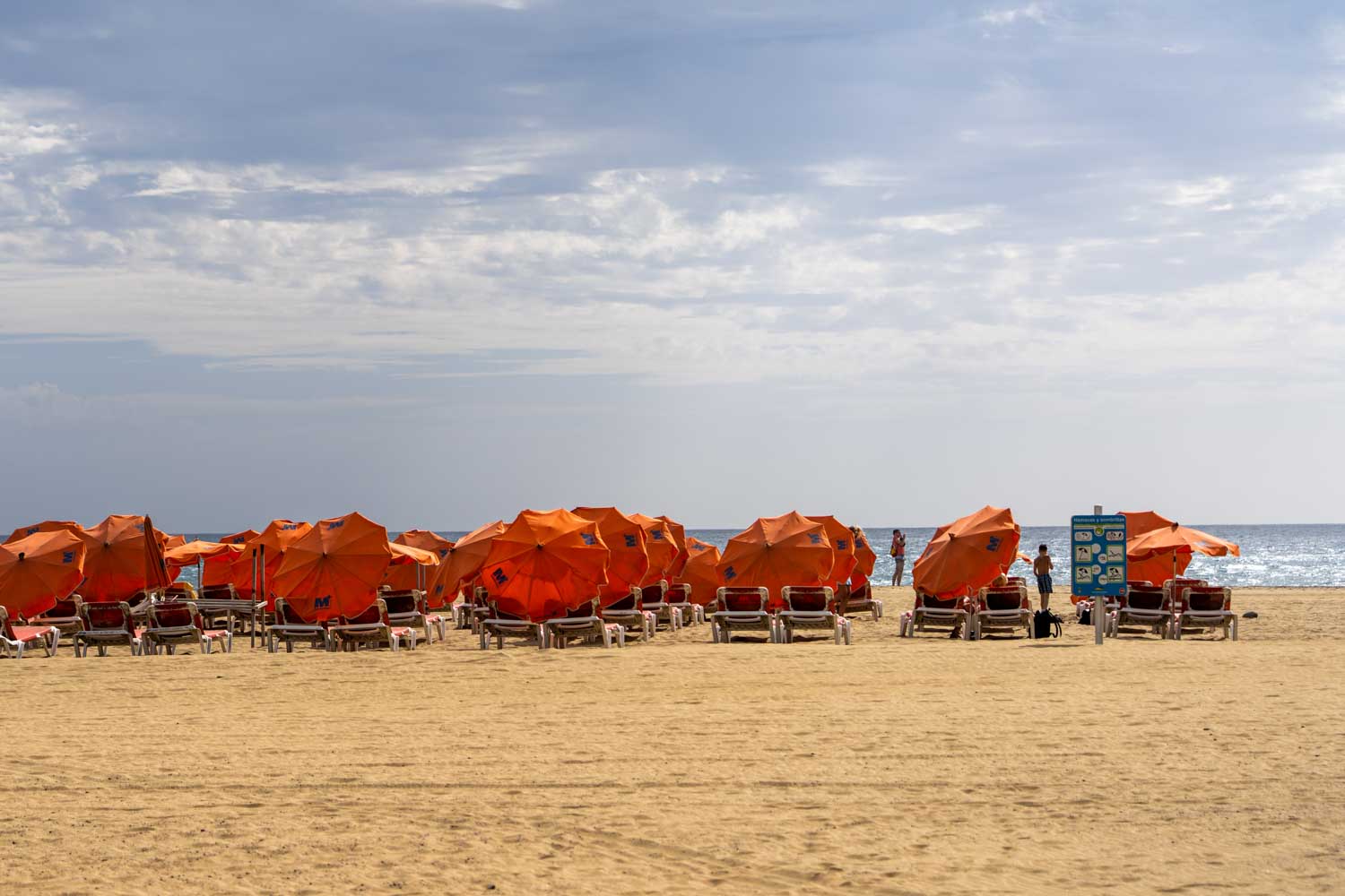 Orange beach umbrellas and loungers on a sandy shore under a cloudy sky, with people enjoying the sea in the distance.