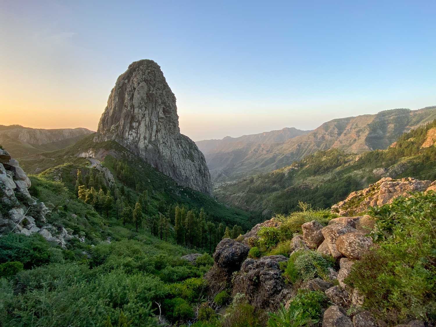 Rocky peak against a sunrise in a lush, green mountain landscape.
