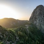 Sunrise behind a rocky mountain peak with lush greenery and winding road in foreground.