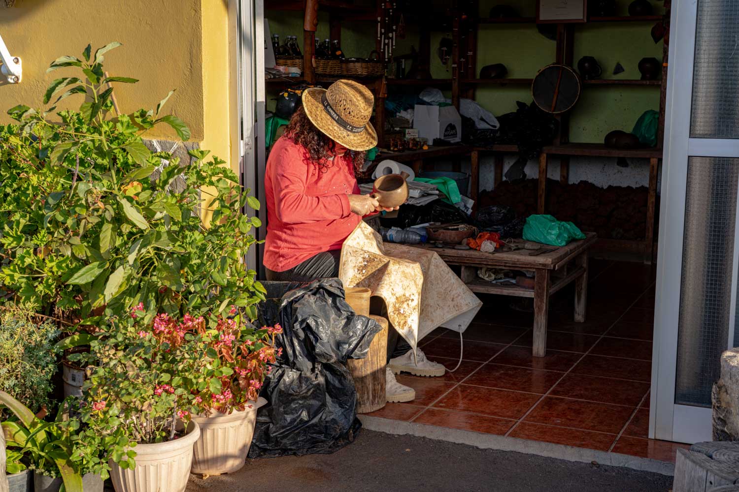 Person in red shirt and hat crafting pottery outside a shop, surrounded by plants and pottery supplies.
