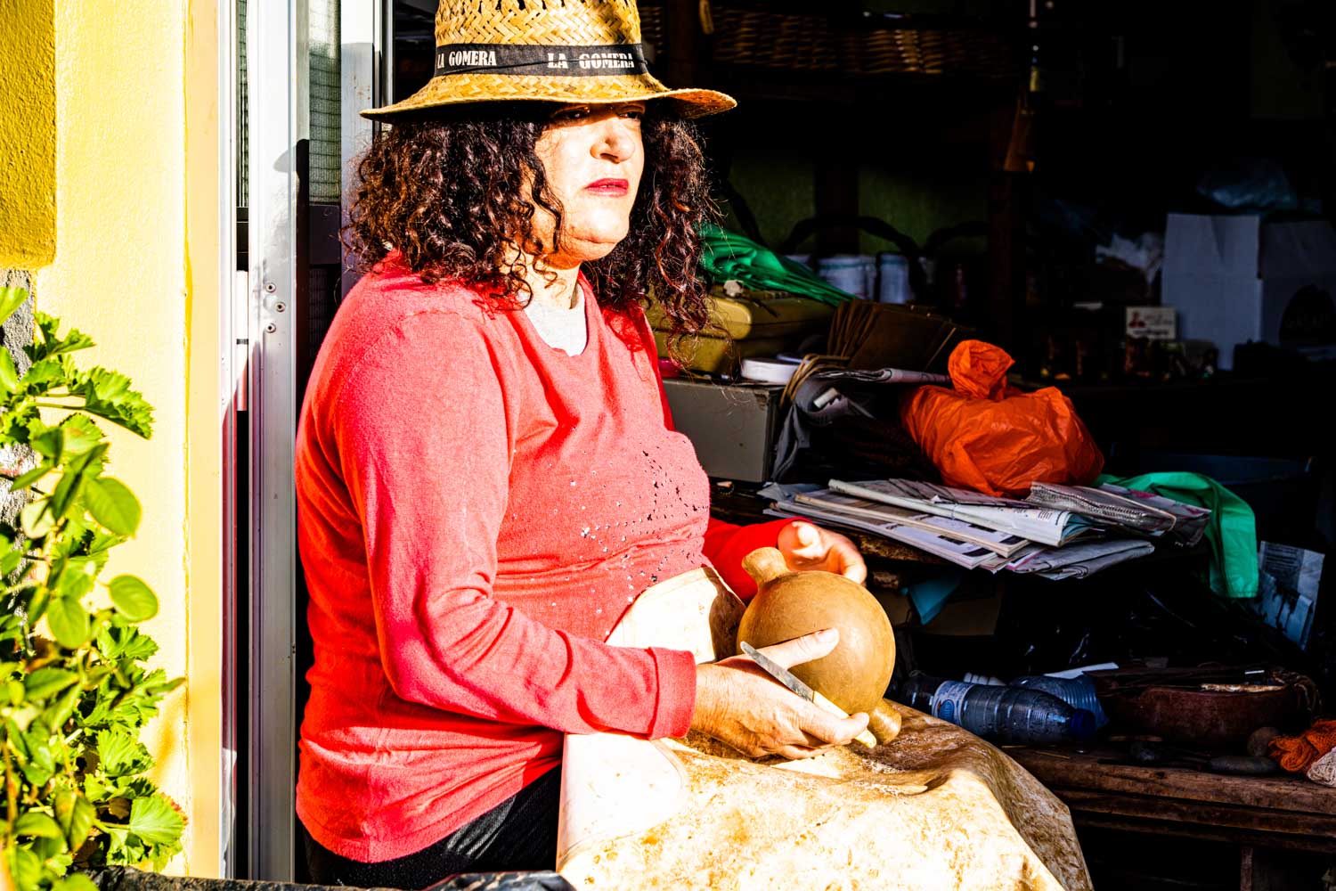 Woman in a straw hat crafts pottery outdoors, wearing a red shirt, with plants and supplies around.