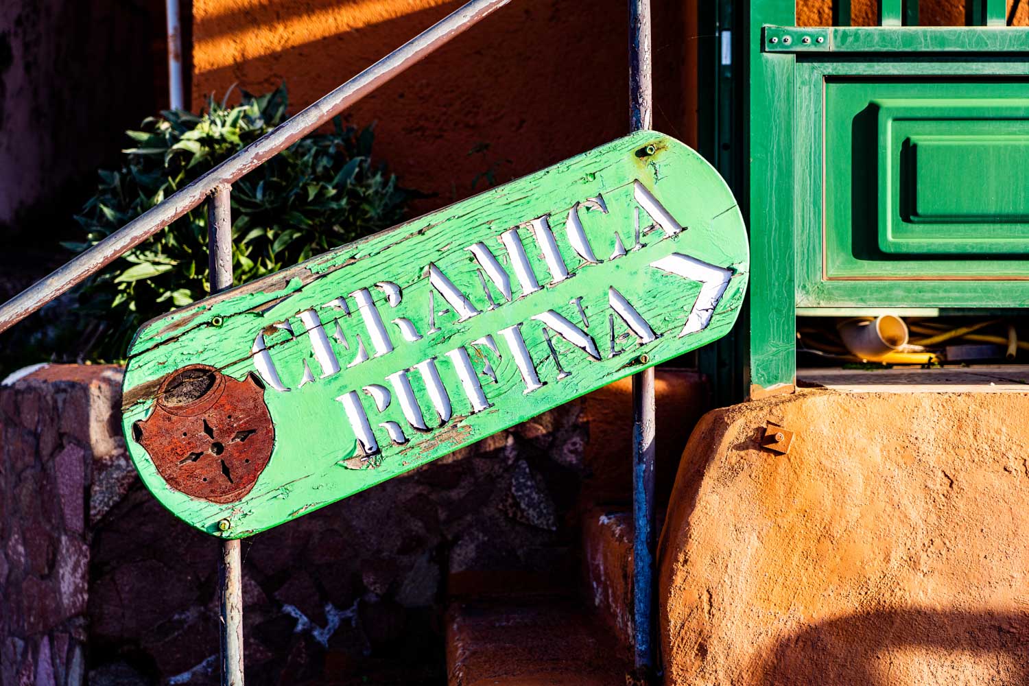 Green sign reading Ceramica Rufina with pottery icon, mounted on rustic orange wall next to green door.
