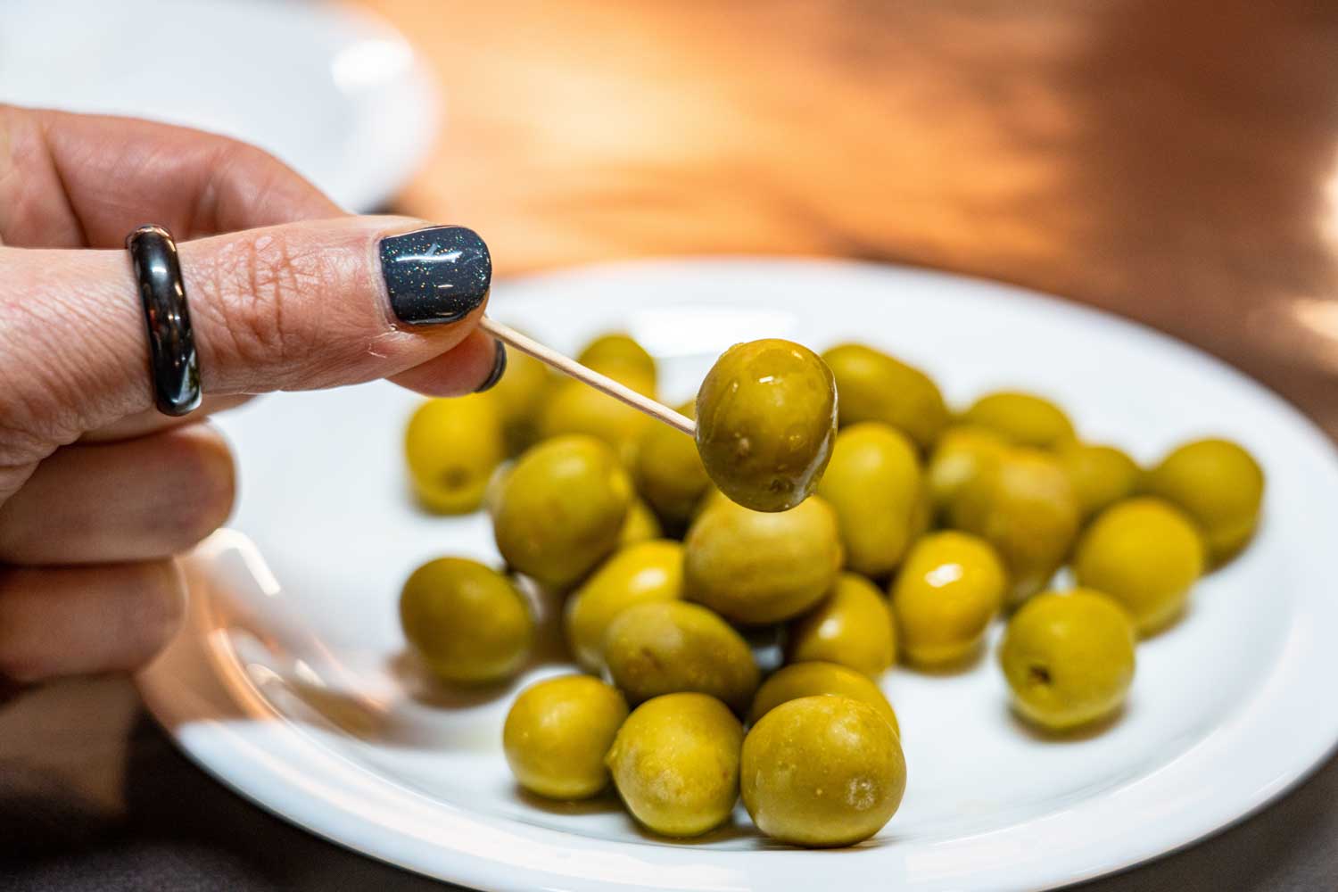 Hand with black nail polish holds green olive on a toothpick over white plate with more olives.