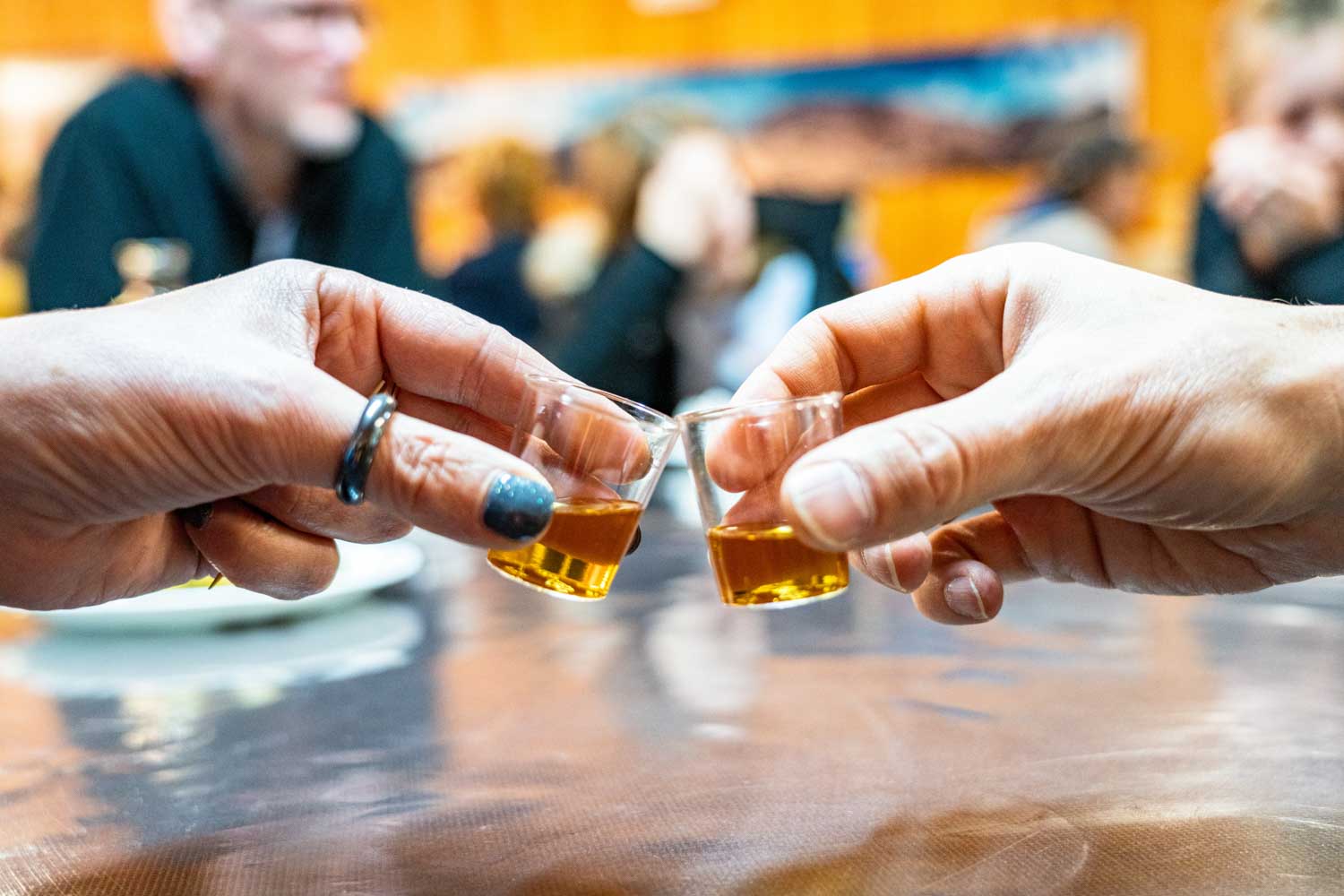 Hands clinking small glasses of whiskey in a social setting with blurred people in the background.