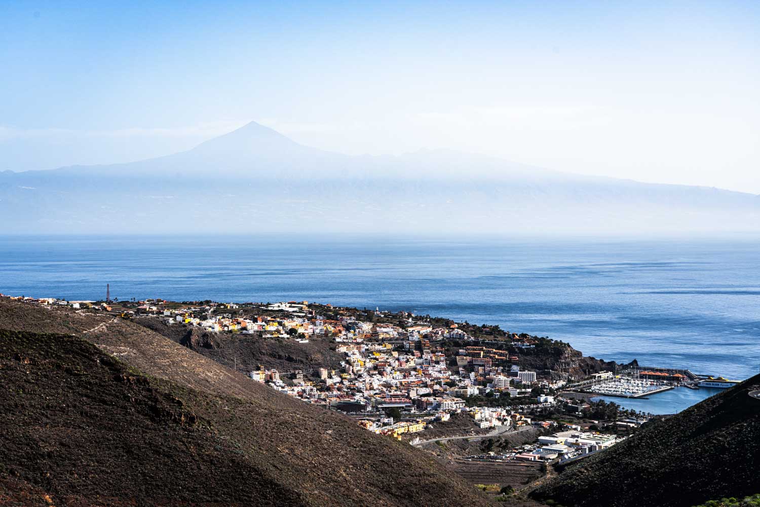 Coastal town with colorful buildings and marina bordered by lush hills and distant mountains under a clear blue sky.