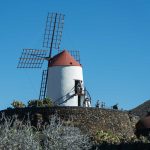 Historic windmill with red roof and tourists on stone terrace against blue sky.