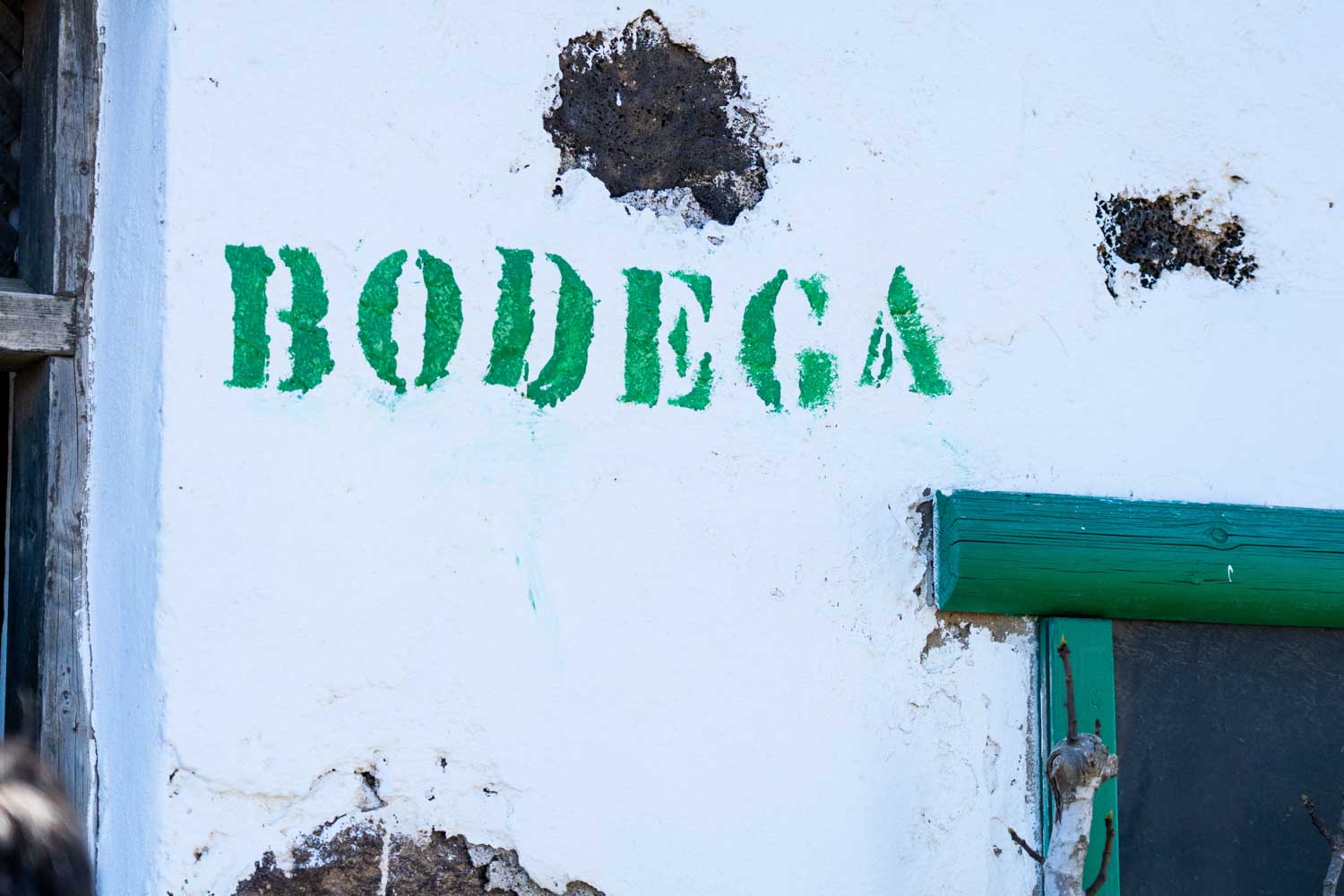 White wall with green 'Bodega' text and weathered patches, adjacent to a window frame on a rustic building exterior.