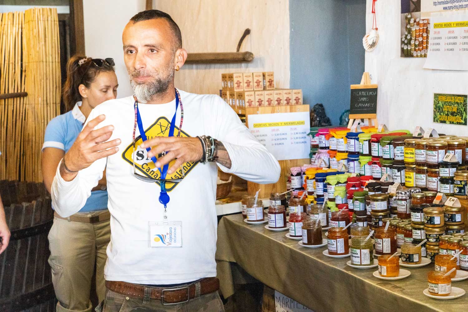 Man explaining products at a jam and sauce stall, colorful jars displayed, indoor market setting.
