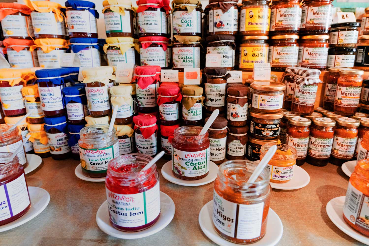 Assorted colorful jam jars on a display table with open jars and sample spoons in a market setting.
