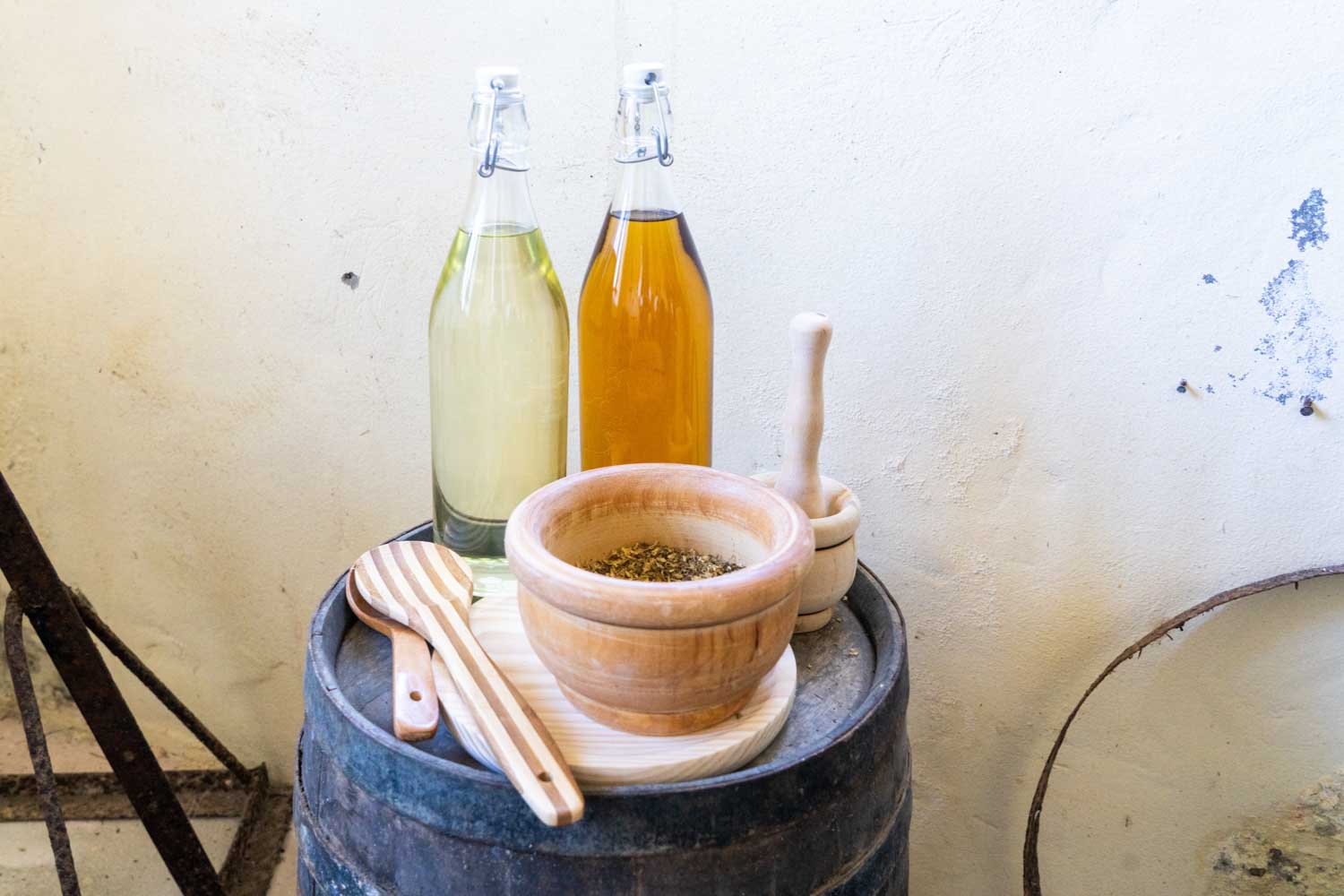 Wooden mortar and pestle, two bottles of oil on a barrel with wooden utensils against a rustic white wall background.